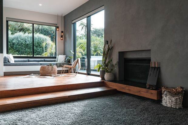 Contemporary living room with a sunken seating area, broad dark-framed glazing, a built-in fireplace set into a textured grey wall, and warm timber steps and hearth ledge. The room includes a low window seat, lounge chairs, indoor plants, woven accessories, and a large charcoal rug, creating a refined indoor-outdoor connection.