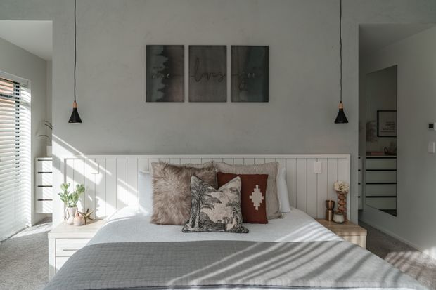 A bright, softly styled bedroom with a bed centred against a white vertical-panelled feature wall. The bed is layered with neutral grey bedding and textured cushions, flanked by white bedside tables, while black pendant lights hang on either side. Natural light enters through a window with white horizontal blinds, and framed artwork hangs above the bed. A doorway and built-in storage are visible to the right.