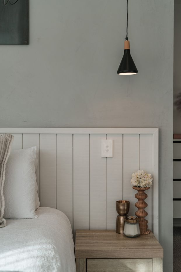A softly styled bedroom vignette featuring a partially visible bed with white bedding, a vertical-panelled light wall headboard, and a wood bedside table. A black pendant light with a timber accent hangs above, while the bedside is decorated with a ribbed metallic cup, ceramic vessel, and sculptural wooden vase holding pale flowers. The palette is muted and neutral with warm timber details.