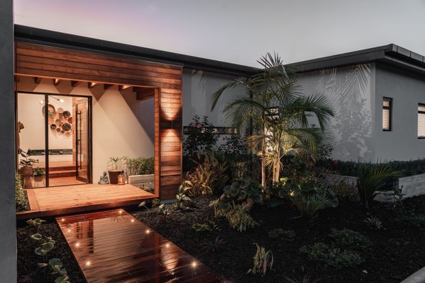 A dusk view of a modern single-storey home arranged around a landscaped courtyard. The entrance is framed in warm horizontal timber cladding, with full-height glazed doors opening onto a timber deck and a reflective timber walkway with integrated lights. Dark charcoal roof edging and smooth grey rendered walls contrast with lush tropical planting and a palm tree. Warm interior and exterior lighting create an inviting atmosphere.