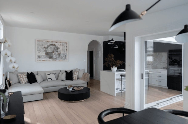 Bright open-plan living and kitchen interior with pale timber flooring, white walls, and a light grey sectional sofa arranged around a low round black coffee table. A black dining table and chairs occupy the foreground, while a white kitchen island and black-and-white cabinetry are visible through the wide opening. Large internal glazing, arched doorways, pendant lights, framed artwork, and restrained monochrome furnishings create a refined contemporary setting.