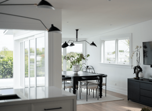 Bright, refined open-plan dining area with a black timber dining table and upholstered chairs, surrounded by large white-framed windows and glazed doors. A sculptural black multi-arm pendant light hangs above the table, while a black sideboard and potted plant add contrast. Pale timber flooring, white walls, and abundant daylight create an airy contemporary interior.