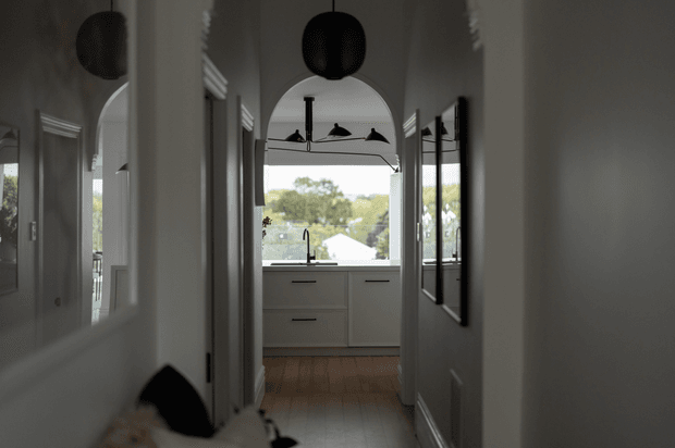 A narrow, softly lit residential hallway leading into a compact kitchen. The corridor has pale walls, timber flooring, arched openings, framed wall mirrors, and dark spherical pendant lights. At the end, the kitchen features light cabinetry, a dark benchtop, a sink and tap, and a large window overlooking leafy surroundings.