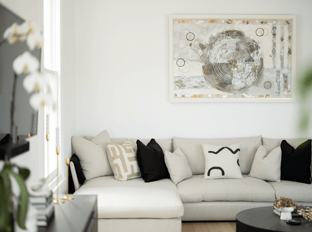 Bright, softly styled contemporary living room featuring a large cream sectional sofa with black and neutral accent cushions, a round black coffee table, pale timber flooring, white walls, and an abstract framed artwork above the sofa. A tall window and indoor greenery add natural light and softness.