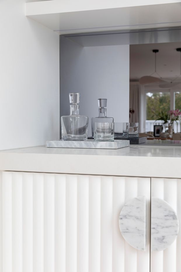 Close-up of a refined white built-in bar or drinks cabinet. The cabinetry has vertical fluted detailing, a white stone or marble benchtop, and distinctive round marble door pulls. A mirrored splashback reflects a bright adjoining living area with pendant lights and glazing. Crystal decanters and glasses sit on a rectangular stone tray, creating a clean, luxurious composition.