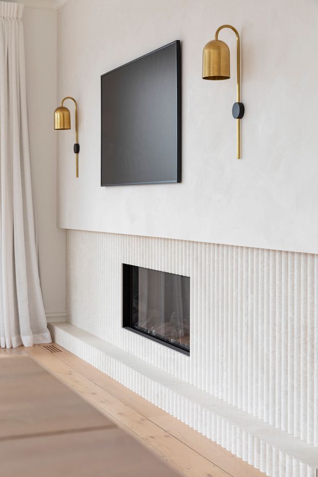 A refined living room feature wall with a large wall-mounted black television, symmetrical brass wall sconces, and a linear fireplace set into a white vertically fluted surround. Full-height cream curtains, pale timber flooring, and a neutral upholstered seating area create a soft, minimalist interior.