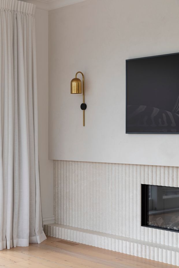 Close-up of a refined living room media and fireplace wall. A wall-mounted television sits above a built-in linear fireplace, both integrated into a pale textured fluted wall treatment with a projecting mantel-like ledge. A slim brass wall sconce provides a warm accent, while full-height off-white curtains frame the left side and light timber flooring grounds the space.