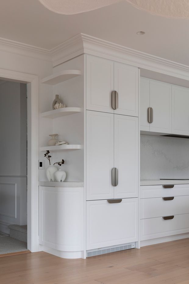 Bright, refined white built-in kitchen cabinetry with integrated appliances, framed shaker-style doors, brushed metal pulls, open curved display shelving, and a pale textured backsplash. The cabinetry is set against warm light timber flooring and detailed white crown moulding, with a doorway and carpeted stair area at left.