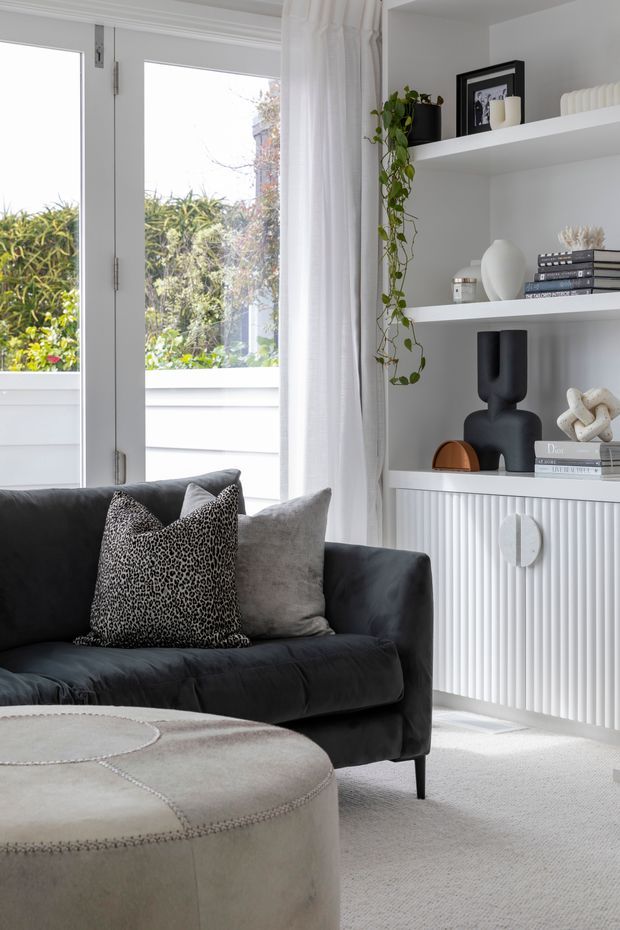 Bright, refined living room corner with a deep charcoal sofa, neutral upholstered ottoman, layered cushions, white sheer curtains, and built-in white shelving. The shelving features ribbed wall panelling, decorative objects, books, framed artwork, and trailing greenery. Full-height glazed doors open toward a leafy garden, bringing in abundant natural light.