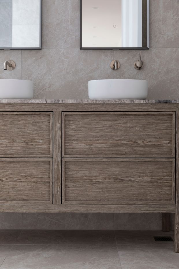 A refined contemporary bathroom vanity with two white vessel basins, brushed metal wall-mounted taps, large rectangular mirrors, and a suspended timber vanity with integrated drawers. The walls and floor are finished in large-format stone-look tiles, creating a muted, luxurious neutral scheme.