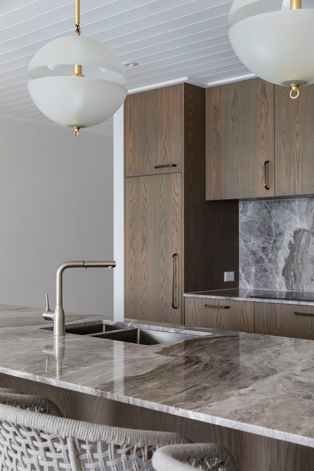 A refined kitchen interior featuring dark natural-wood cabinetry, a large island with a dramatic veined stone benchtop and integrated double sink, brushed metal tapware, and a matching stone splashback. White vertical-panel ceiling boards and sculptural white-and-brass pendant lights add contrast, while woven bar stools sit at the island.
