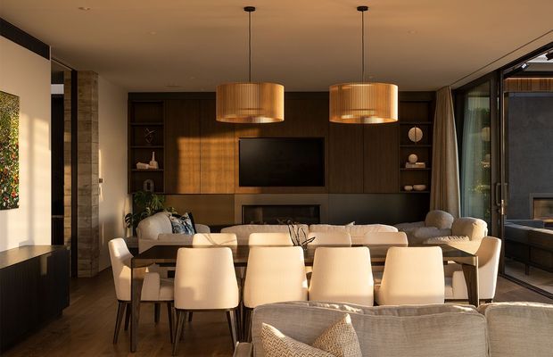 Warm, sophisticated open-plan living and dining area with dark timber built-in cabinetry, a wall-mounted television, a large dining table with upholstered chairs, pendant lighting, and a neutral sectional sofa. Full-height glazing on the right opens toward an outdoor space, while layered timber and soft furnishings create an inviting contemporary interior.