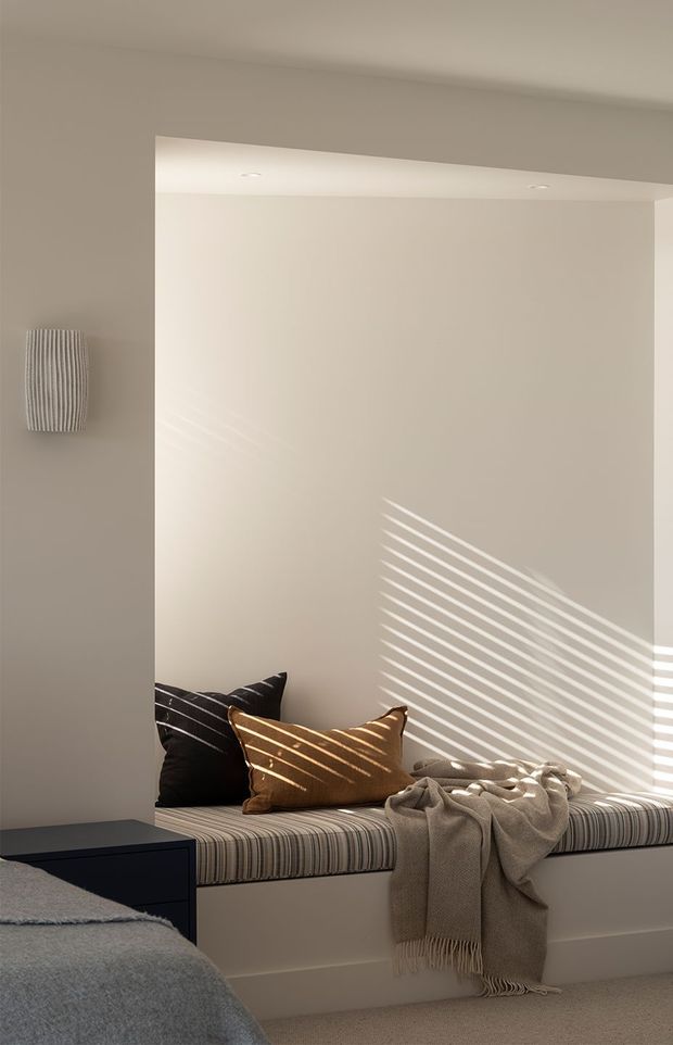 A softly lit bedroom featuring a built-in window seat with striped upholstery, dark and rust-toned cushions, and a draped neutral throw. A bed is partially visible in the foreground, while diagonal sunlight filtered through blinds creates strong linear shadows across the pale walls and cushions. The room has a calm, warm, minimalist appearance with recessed ceiling lighting and neutral finishes.