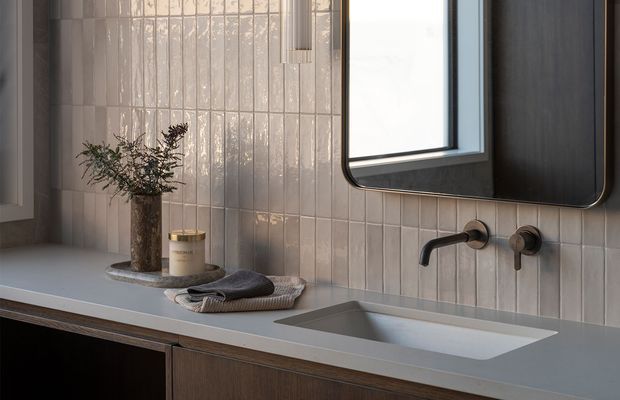Close-up view of a refined contemporary bathroom vanity. A pale integrated benchtop contains a rectangular inset basin, set above dark timber cabinetry. The wall is finished in vertically stacked glossy pale tiles, with a wall-mounted brushed dark-metal tap and a rounded rectangular mirror. A small vase and folded towels add subtle styling.