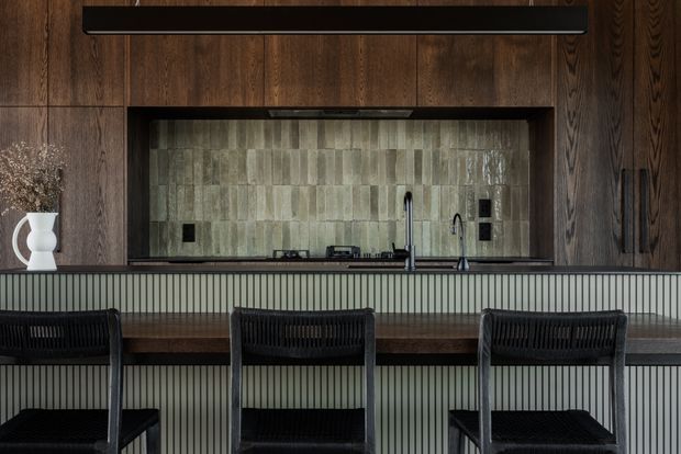 A sophisticated kitchen and dining counter featuring dark vertical timber wall panelling, a large framed green-grey tiled splashback, dark cabinetry, and a fluted light-grey island or breakfast bar. Three black woven bar stools line the timber counter, with a black linear pendant above. The space has a moody, refined contemporary feel with strong textural contrast.