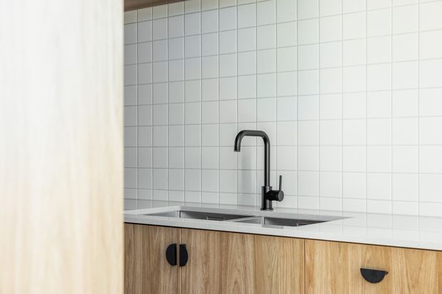 A compact modern kitchen sink area viewed partially behind a light timber partition. The scene features white square tiled walls, a stainless-steel double-bowl sink, a tall dark metal gooseneck tap, and warm wood-grain base cabinetry with dark half-moon handles.