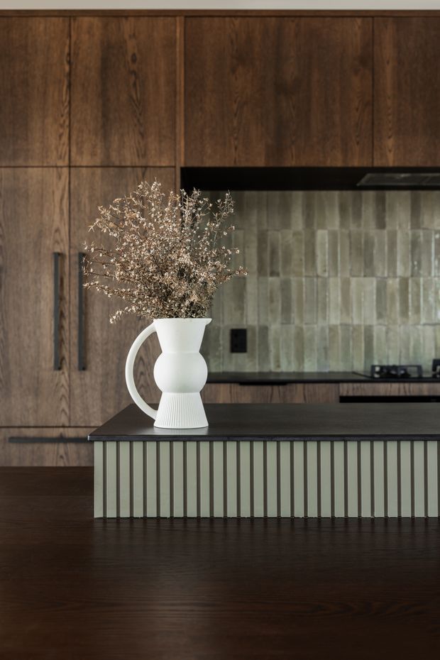 A refined kitchen interior featuring extensive dark timber cabinetry, a dark benchtop, and a muted green-grey vertically fluted island front. The backsplash is composed of textured rectangular tiles in soft grey-green tones, with integrated black appliances and hardware. A white sculptural ceramic vase with dried branches sits on the island. The composition is warm, moody, and contemporary with strong natural material contrast.