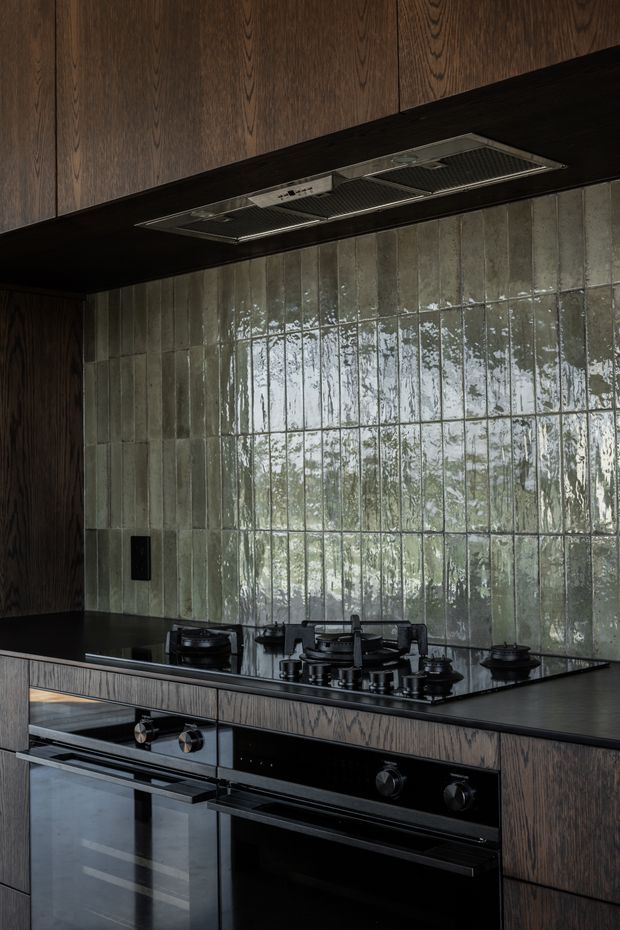 Close-up view of a sophisticated dark-toned kitchen featuring vertical dark timber cabinetry, a black stone benchtop, integrated black cooking appliances, and a large glossy textured green-grey tiled splashback. A concealed stainless-steel rangehood is set beneath the overhead cabinetry.