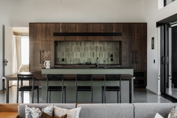 Contemporary open-plan living and kitchen area centred on a large island with integrated seating. The room features dark timber full-height cabinetry, a fluted island front, a reflective grey-green tiled splashback, dark stone-look flooring, black-framed glazing and a grey sectional sofa in the foreground. A dining area and bright curtained opening are visible to the left.