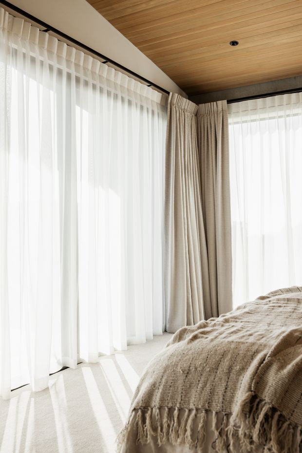Bright, softly styled bedroom corner with floor-to-ceiling sheer curtains and beige drapery wrapping around large windows. A textured neutral throw covers the edge of the bed, while the warm timber-lined ceiling and pale carpet create a calm, natural atmosphere. Recessed ceiling lighting and dark curtain tracks provide subtle contrast.