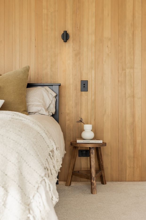A warmly styled bedroom corner featuring a bed with layered cream bedding and a tan cushion, set against a full-height vertical timber-panelled feature wall. A small rustic timber stool serves as a bedside table with a ceramic vase and books, while dark electrical outlets and a small wall-mounted light provide functional detailing. Neutral carpet and natural wood tones create a calm, earthy atmosphere.