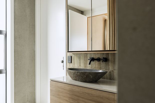 Compact contemporary bathroom vanity area featuring a dark natural stone vessel basin on a light stone benchtop, timber-grain cabinetry, textured neutral wall tiles, black wall-mounted tapware, and mirrored storage above. A white door and partial window are visible at the side.
