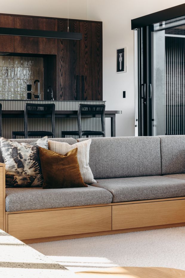 A refined open-plan living room with a large grey upholstered sofa in the foreground, featuring a warm timber plinth and built-in storage drawers. Behind it is a compact kitchen with dark stained timber cabinetry, a dark benchtop, and integrated pendant lighting. Black-framed glazed doors bring in strong natural light, while neutral walls, a textured pale rug, and earthy cushions create a warm, contemporary atmosphere.