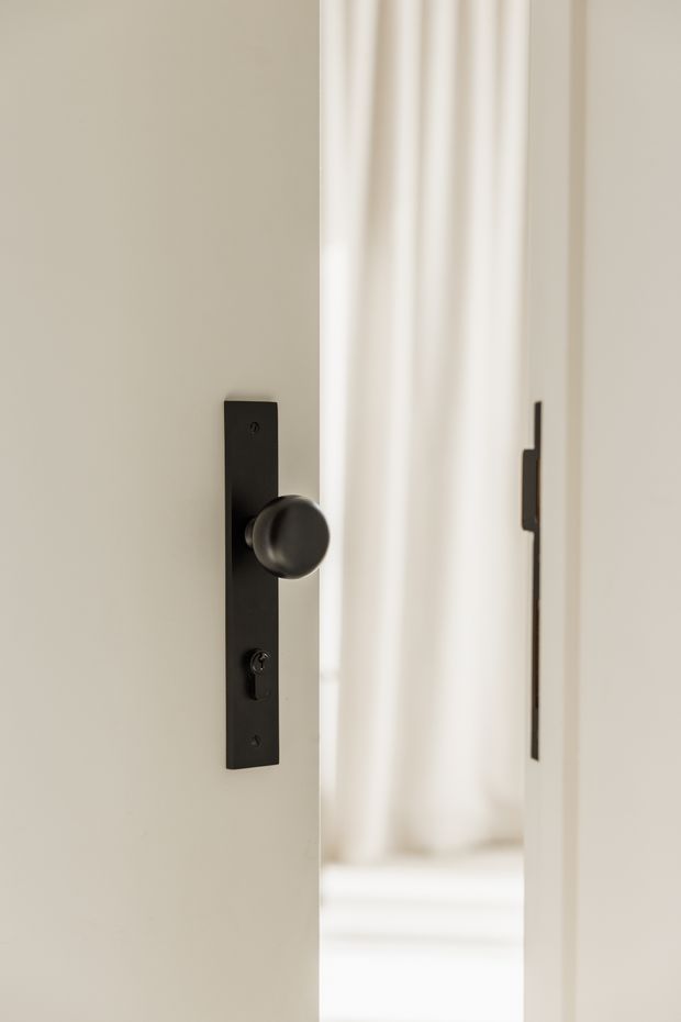 Close-up of a partially open interior door featuring a matte black rectangular backplate, round black knob, and keyhole escutcheon. The surrounding walls and door surfaces are warm off-white, with a softly lit bright room visible through the opening.