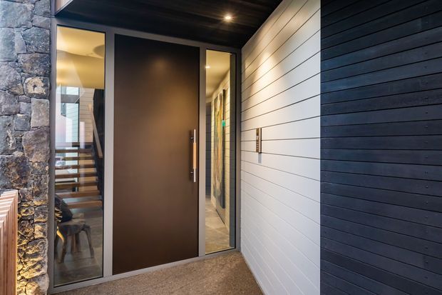 Contemporary residential entry viewed from outside, featuring a tall dark pivot door with narrow glazed sidelights, horizontal light and dark wall cladding, a stone-clad wall, recessed ceiling lighting, and a glimpse of a timber staircase and interior beyond.