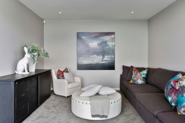 A compact, softly styled living room with a dark sectional sofa, white accent armchair, round upholstered ottoman, grey carpet, neutral walls, recessed ceiling lights, and a dark timber sideboard. Colourful cushions, greenery, and a landscape artwork add decorative accents.