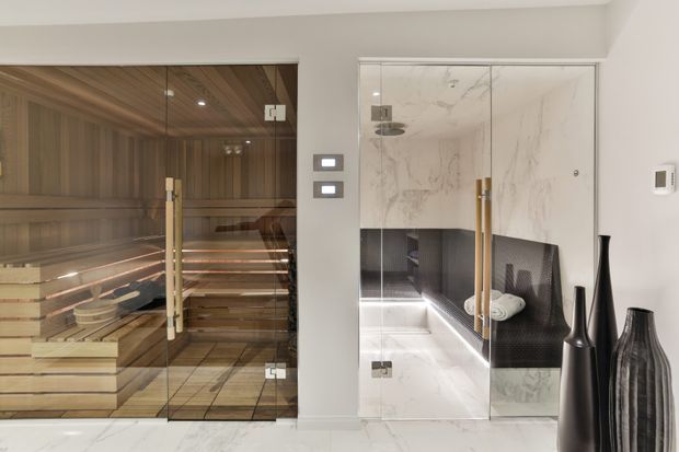 A refined spa-style interior featuring a glass-enclosed timber sauna on the left and a glass-enclosed marble-lined steam room or shower area on the right. The space has white marble flooring and walls, warm timber sauna cladding and benches, dark reflective wall surfaces, integrated lighting, and sculptural black vases.