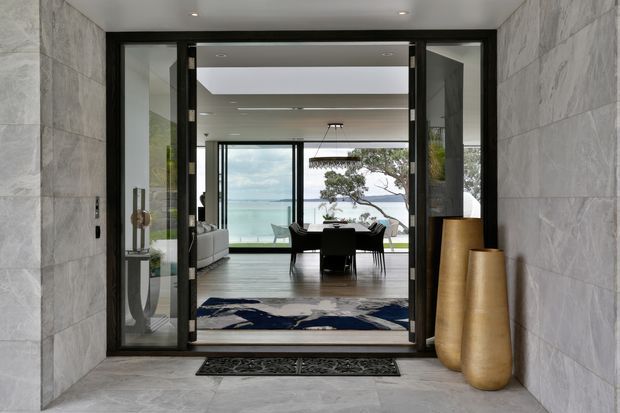 A refined modern entryway with large-format grey stone wall and floor finishes, black-framed glazed double doors, and tall sculptural gold-toned vases. Through the entrance is an open-plan living and dining area with a contemporary pendant light, dark dining furniture, pale seating, and expansive glazing overlooking a coastal waterside setting.