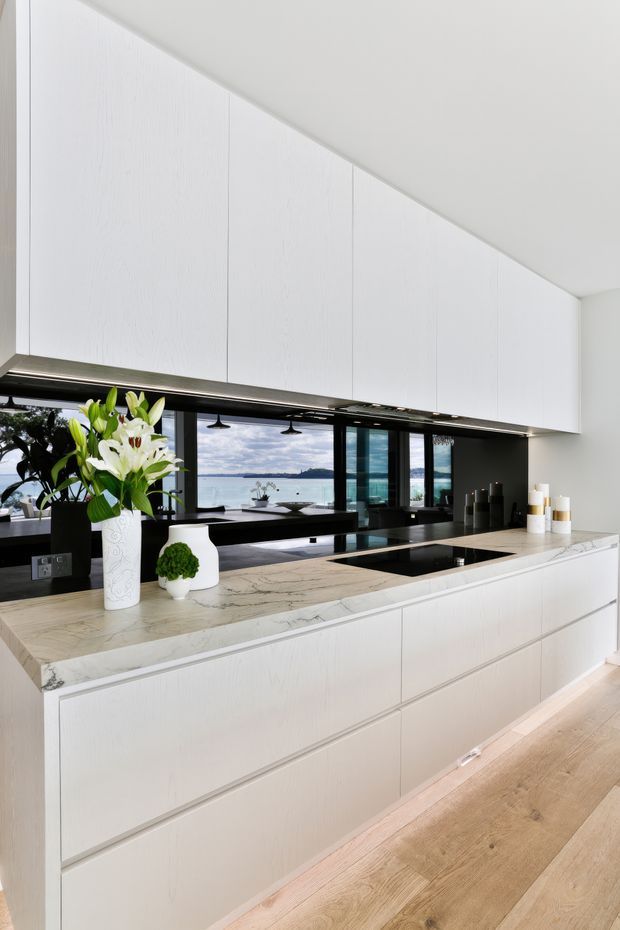 Bright contemporary kitchen with extensive white handleless cabinetry, a large pale veined stone island benchtop, integrated black cooktop and sink, dark window framing, and light timber flooring. Floor-to-ceiling glazing behind the kitchen offers a waterfront view, while integrated strip lighting illuminates the cabinetry and benchtop.