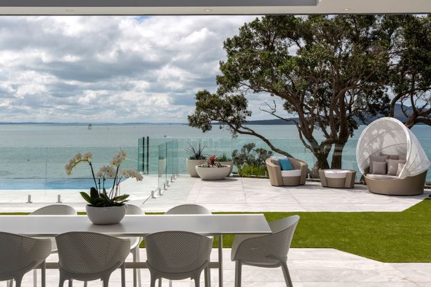 A refined waterfront outdoor entertaining area viewed from beneath a covered roof edge. The foreground has a white dining table with woven outdoor chairs and potted orchids, opening onto a pale tiled terrace, manicured lawn, glass pool or terrace fencing, and a turquoise bay beyond. A mature tree, sculptural planters, lounge chairs, and a rounded outdoor daybed create a resort-like setting.
