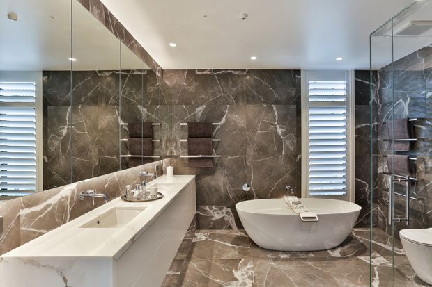 A luxurious contemporary bathroom featuring dramatic dark marble-look wall and floor surfaces, a freestanding white bathtub, long double vanity with integrated basins, mirrored wall, glass-enclosed shower, and plantation-shutter windows. Recessed ceiling lighting creates a polished, refined atmosphere.