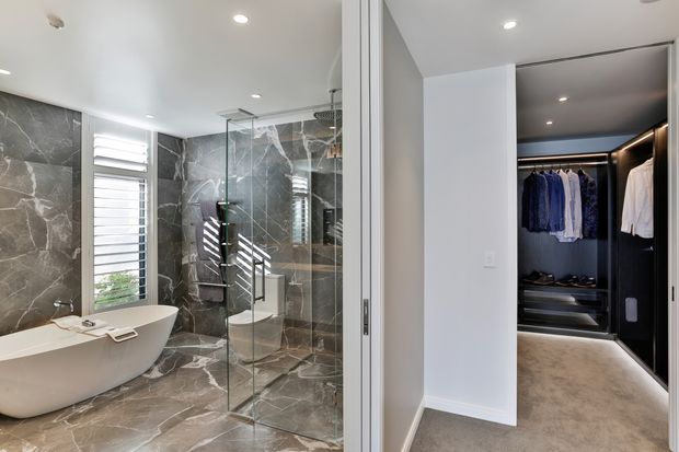 A refined bathroom with large-format dark marble-look wall and floor tiles, a freestanding white bathtub, and a frameless glass shower enclosure. Through the open doorway is a dark, illuminated walk-in wardrobe with hanging clothes and shelving. Recessed ceiling lights and white plantation shutters provide a clean, contemporary finish.