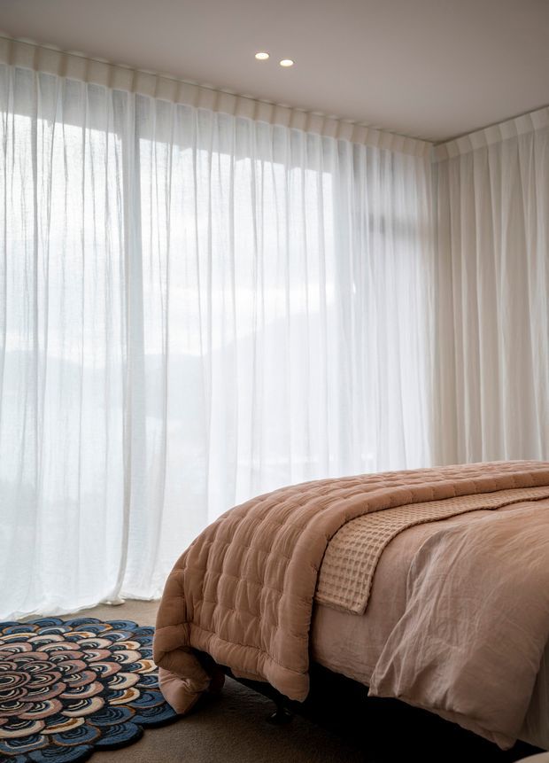 A softly lit bedroom featuring a large bed dressed in layered blush and beige bedding, floor-to-ceiling sheer curtains wrapping around a corner, recessed ceiling lights, and a patterned blue, cream, and terracotta rug. The room has a calm, warm, textural aesthetic.