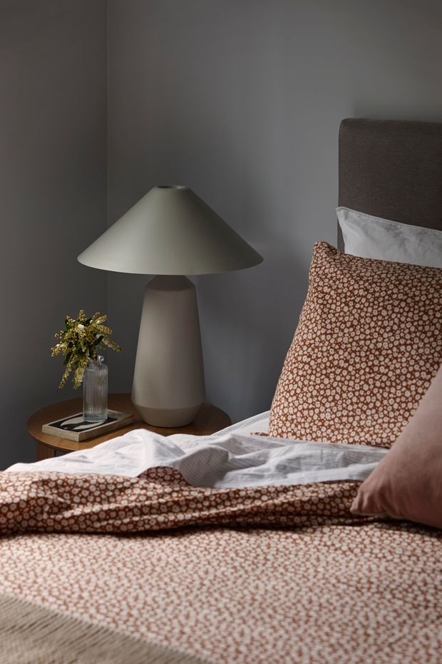 A close-up, warmly styled bedroom scene featuring a bed dressed in a rust-brown and cream patterned duvet and pillows, a dark upholstered headboard, and a rounded ceramic bedside lamp on a timber side table. A small glass vase with greenery and stacked books add subtle detail. The room has dark grey walls and a moody, intimate atmosphere.