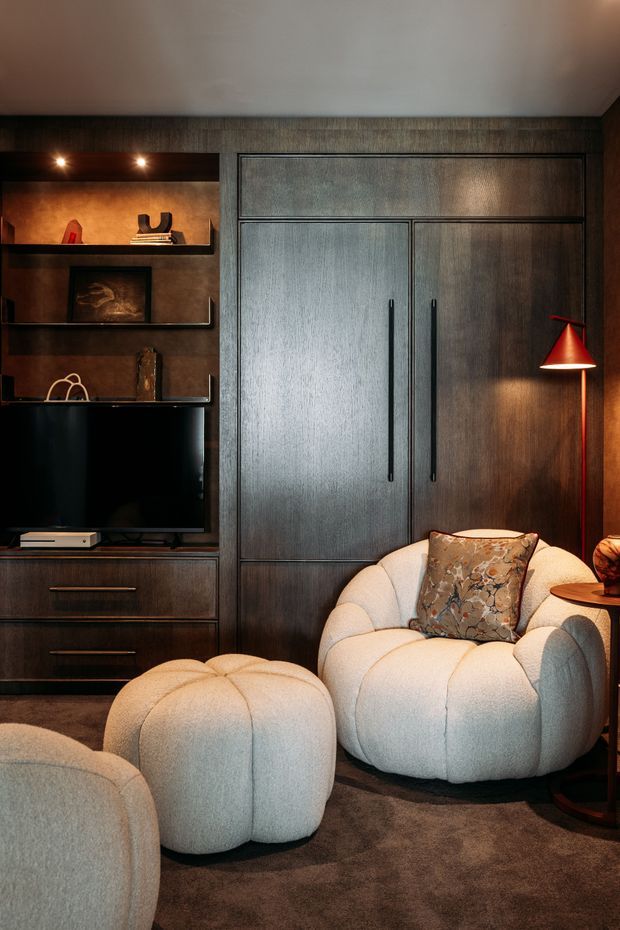 A warm, moody living room featuring dark timber built-in cabinetry with concealed storage, open display shelving, and an integrated television. Plush cream boucle lounge chairs and a matching rounded ottoman sit on a dark carpet, complemented by a red sculptural floor lamp and softly lit decorative shelving.