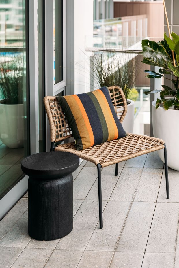 A contemporary balcony or terrace corner with a woven rattan lounge chair, striped cushion, small black sculptural side table, pale tiled flooring, large potted plant, and glazed doors opening toward an urban setting.