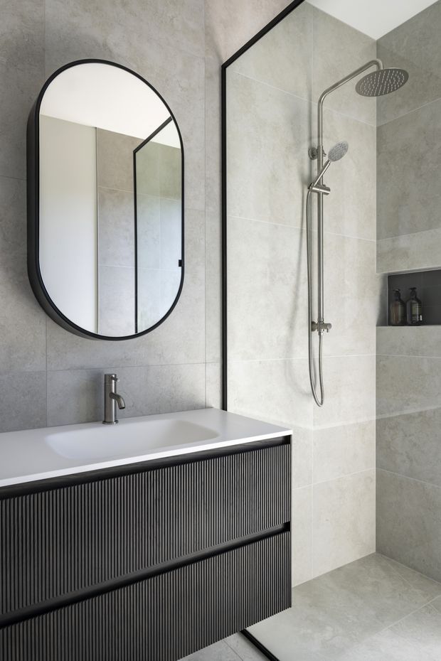 Contemporary bathroom featuring large-format light grey stone-look wall and floor tiles, a frameless glass walk-in shower with overhead and handheld shower fittings, a recessed niche, and a floating dark ribbed vanity with integrated white basin. A tall rounded-rectangle mirror with a black frame adds contrast.