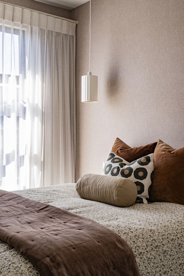 A warm, softly styled bedroom corner with a textured beige wall, full-height layered curtains, a simple white pendant light, and a bed dressed in a muted patterned coverlet. Brown velvet cushions, a cylindrical bolster, and a decorative circular-pattern cushion add tactile contrast and understated character.