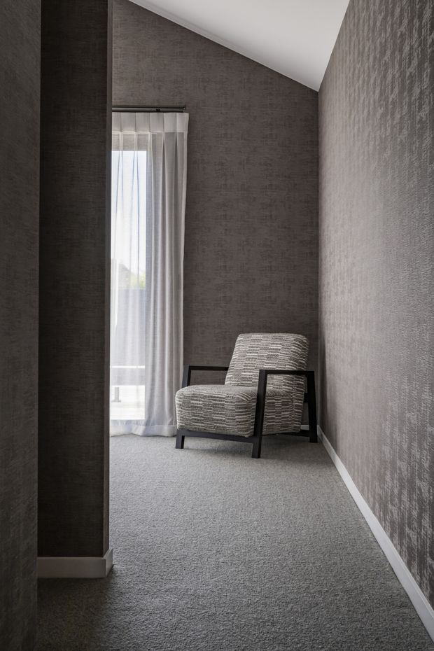 A narrow, tall-ceilinged interior nook or hallway with charcoal-grey textured wall finishes, grey carpet, white skirting boards, and a slim full-height window dressed with sheer curtains. A single upholstered accent chair with a black angular timber or metal frame sits at the far end, creating a quiet monochromatic reading or seating area.