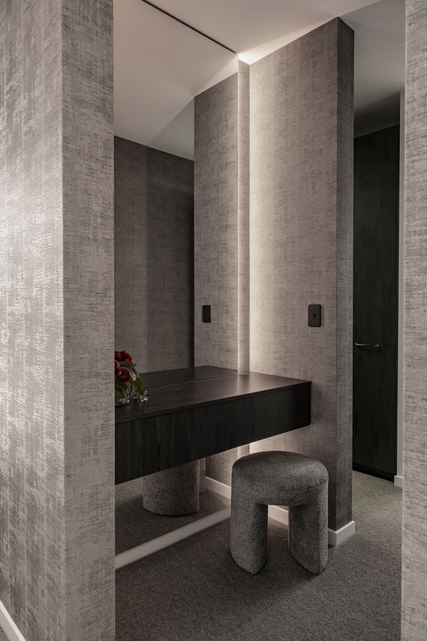 A refined, moody dressing and vanity area with a dark floating timber-look console, integrated mirror, upholstered rounded stool, and softly illuminated textured grey wall panels. A dark doorway and carpeted floor reinforce the subdued, luxurious atmosphere.