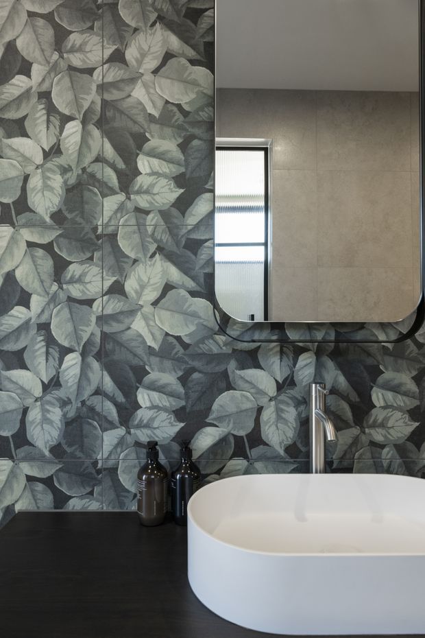 A contemporary bathroom vanity featuring a large white vessel basin on a dark benchtop, a brushed metal tap, and a rounded rectangular mirrored cabinet. The walls are covered in dramatic grey-green botanical leaf-patterned tiles, creating a bold feature backdrop. A narrow frosted window provides natural light, while dark soap dispensers sit beside the basin.