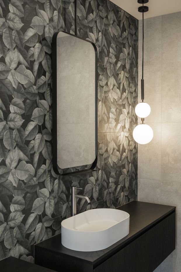 A contemporary bathroom vanity featuring a white vessel basin on a dark fluted built-in cabinet, a black-framed rounded-rectangle mirror, dramatic monochrome botanical wall covering, pale large-format wall tiles, and twin globe pendant lights.