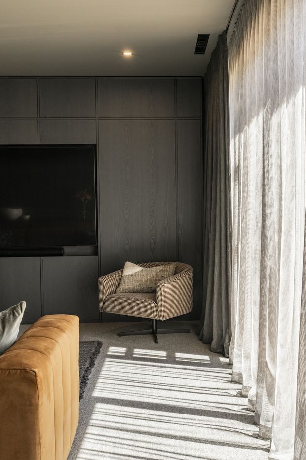 A cozy, contemporary living room corner with a dark full-height timber built-in media wall and integrated television. A tan leather sofa and patterned upholstered armchair sit on a neutral carpet, while full-height sheer curtains and heavier drapes cover a large window, casting strong linear sunlight across the floor.