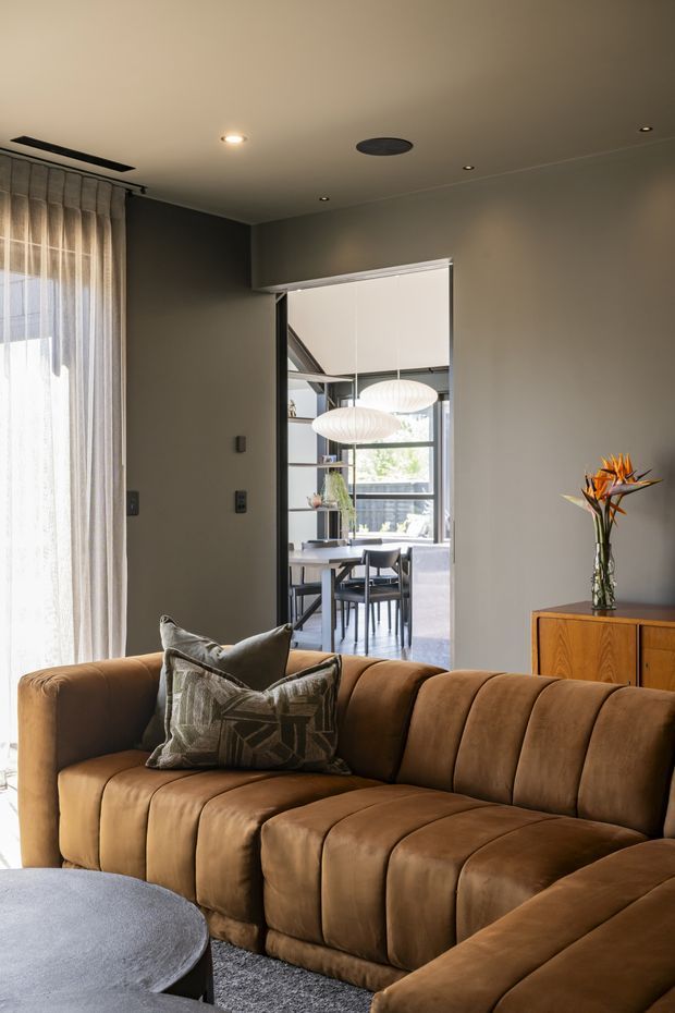 A warm, refined living room centred on a large caramel-brown channel-tufted sectional sofa. The room has dark taupe-grey walls, sheer full-height curtains, a round dark coffee table, grey carpeting, and a timber sideboard topped with a sculptural bird-of-paradise arrangement. A wide opening reveals a bright dining area with a table, chairs, shelving, and pendant lighting. Recessed ceiling lights and a concealed curtain track contribute to the contemporary interior.