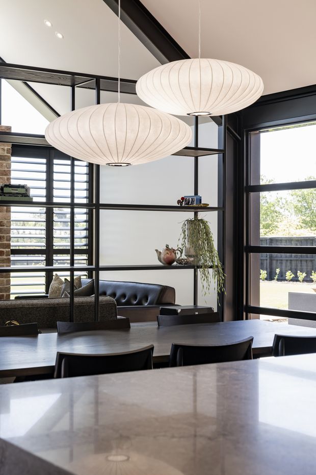A bright, contemporary open-plan dining and living area viewed from a kitchen island or benchtop. The space features a long dark dining table, black-framed steel-look internal glazing and shelving, large louvred windows, exposed dark ceiling beams, a tan leather sofa, and two oversized white sculptural pendant lights. A stone-clad column and indoor plants add texture, while the glazing opens visually toward an outdoor area.