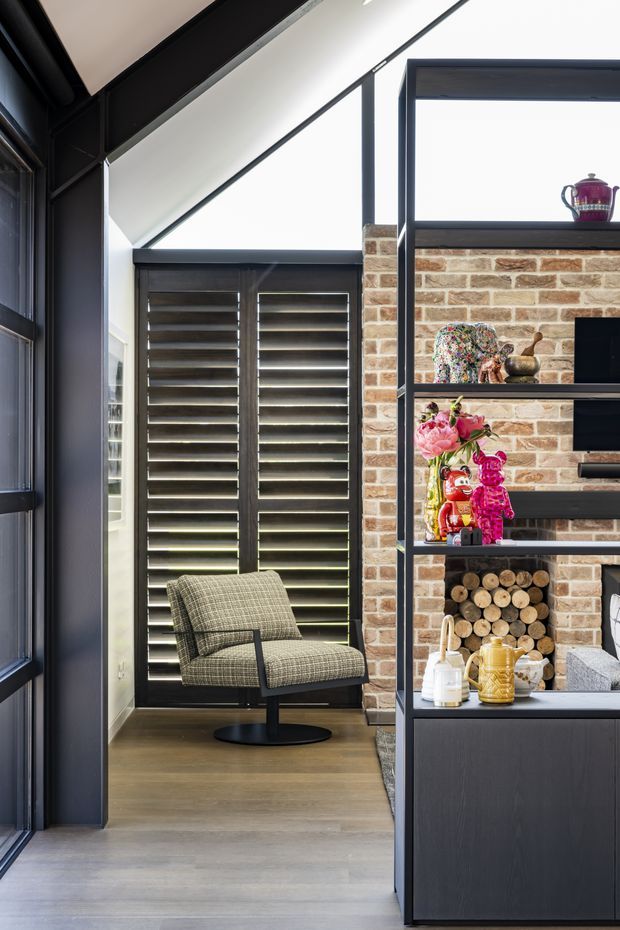 A refined contemporary interior corner featuring a dark-framed glazed partition, black plantation shutters, and an exposed brick feature wall. A woven upholstered lounge chair sits on pale timber flooring beside open black metal shelving displaying decorative objects and stacked firewood. The space has a high, angled ceiling and strong natural light.
